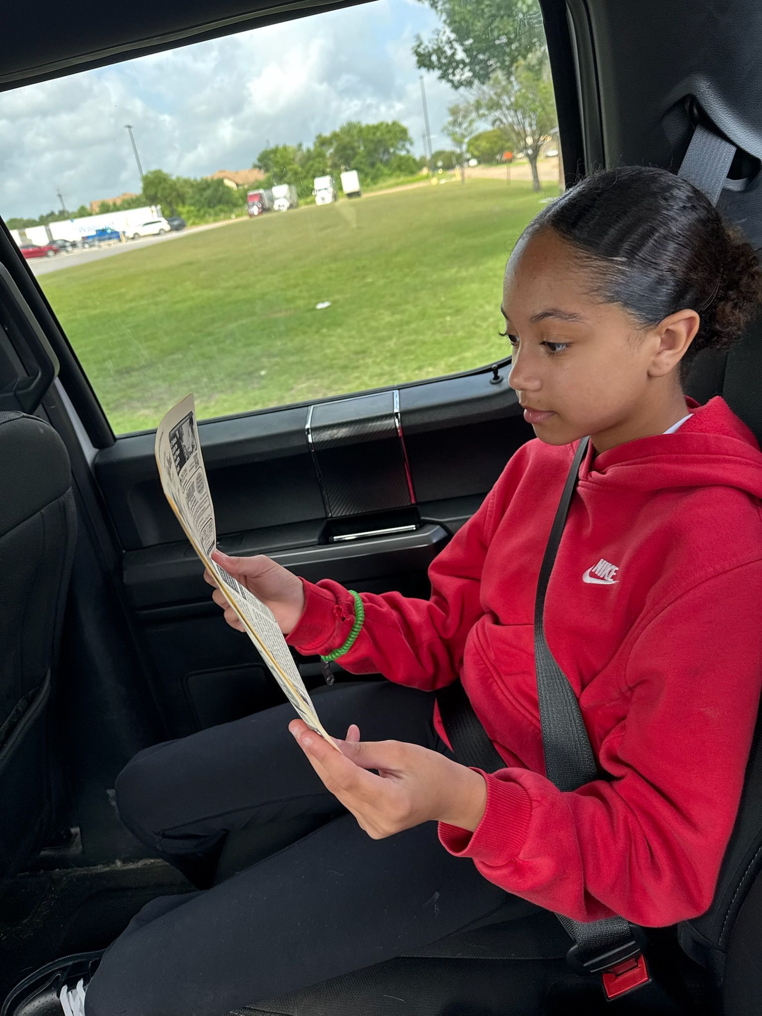 A person in a red hoodie reads a paper inside a vehicle, outdoors visible through the window.