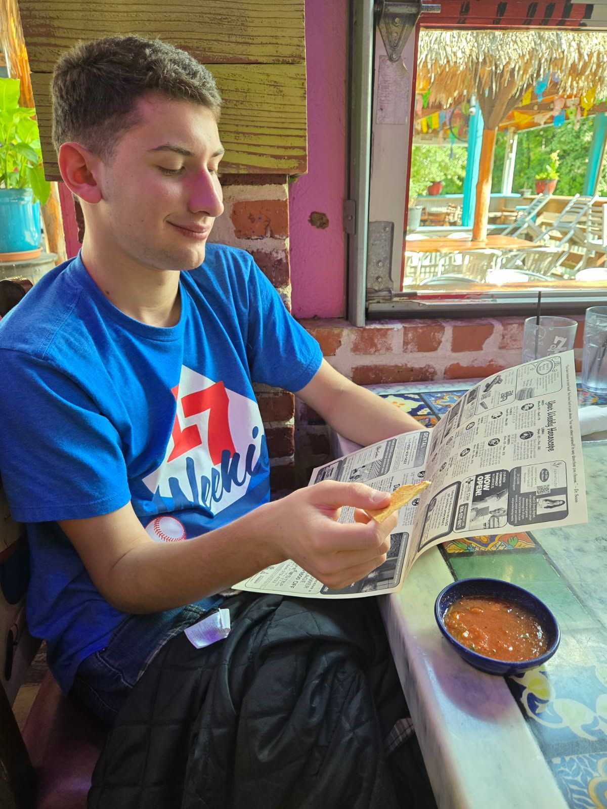 Person in blue shirt reading a menu, holding a chip. Salsa and outdoor seating visible.