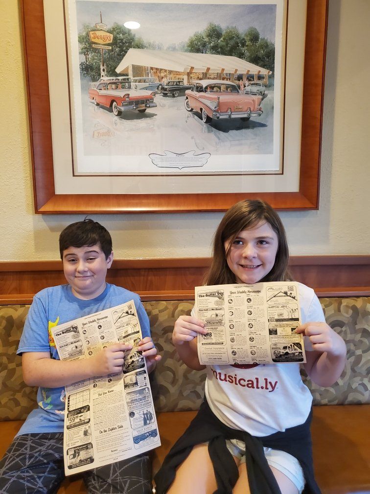 A boy and a girl are sitting on a couch holding newspapers.