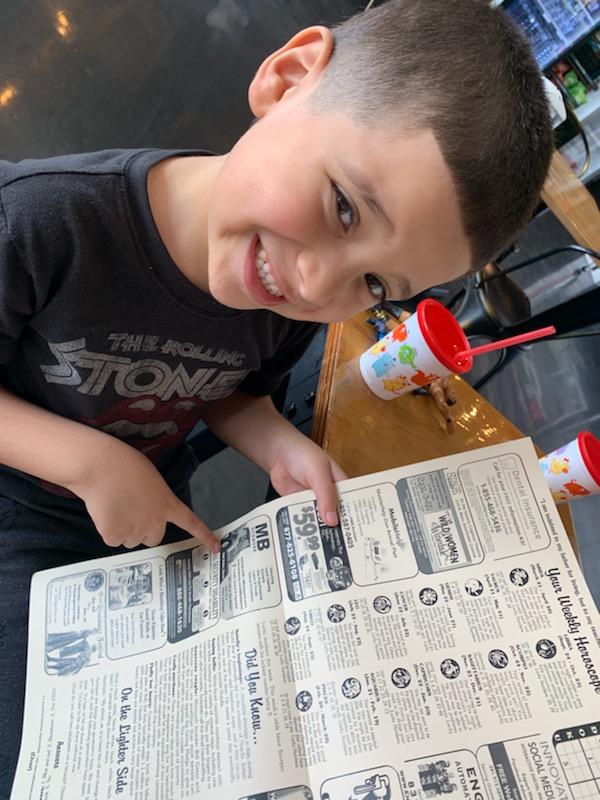 A young boy wearing a tony hawks shirt is reading a newspaper