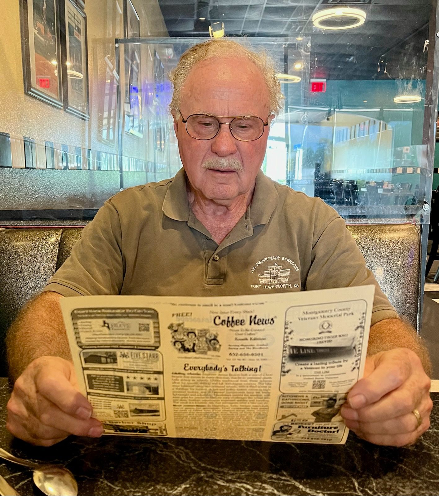Man reading a newspaper at a diner table, wearing glasses. Clear dividers between tables.