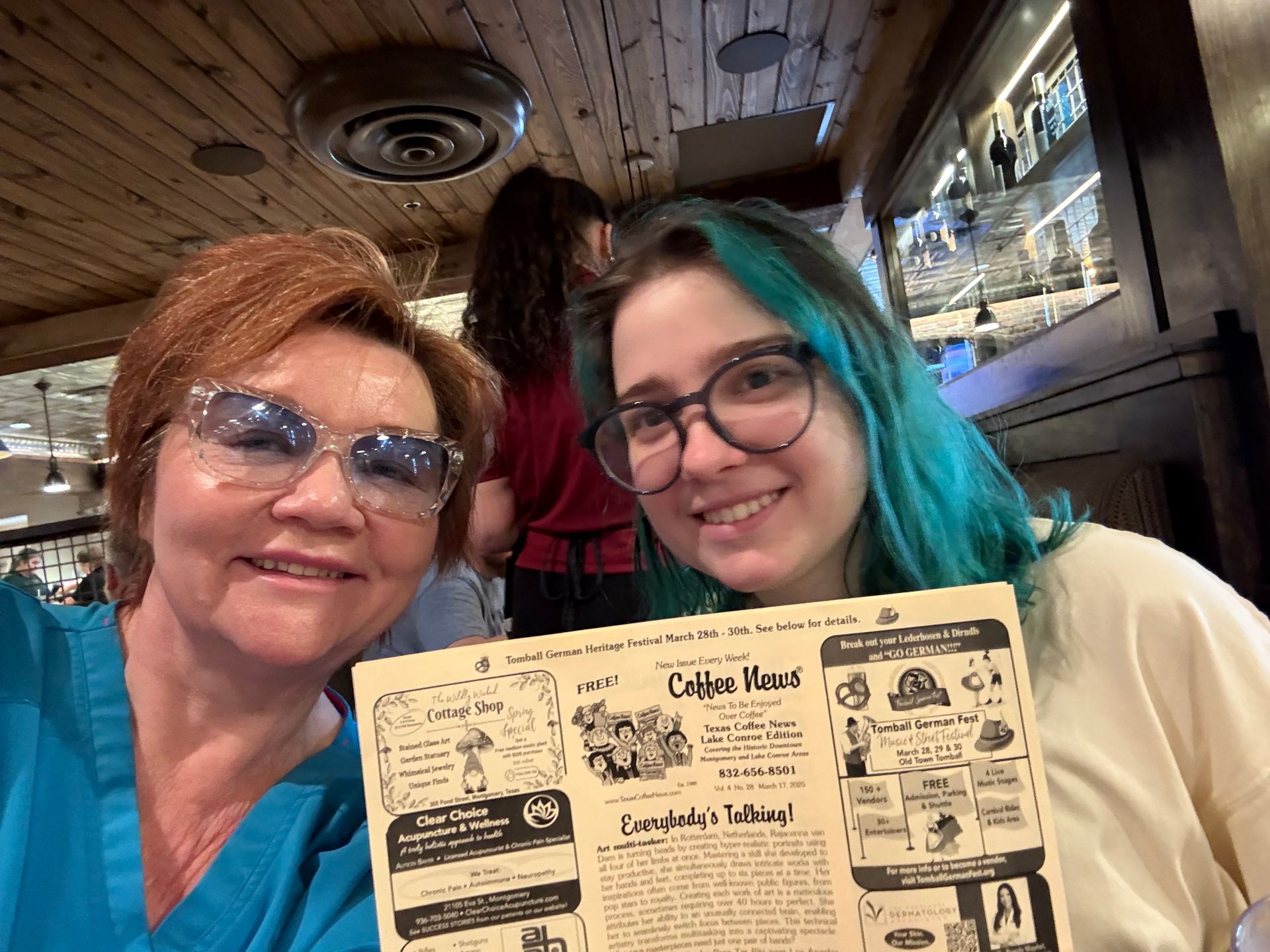 Two people smiling, holding a menu at a restaurant. One with turquoise hair, the other with sunglasses.