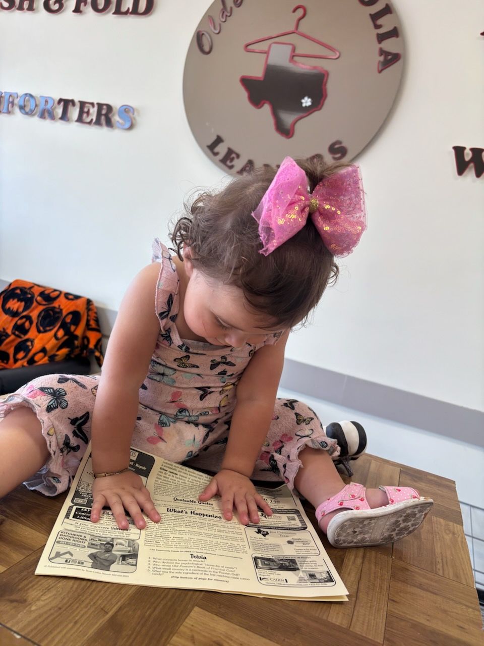 Child with a large pink bow, reading a newspaper on a wooden surface, near a laundromat sign.