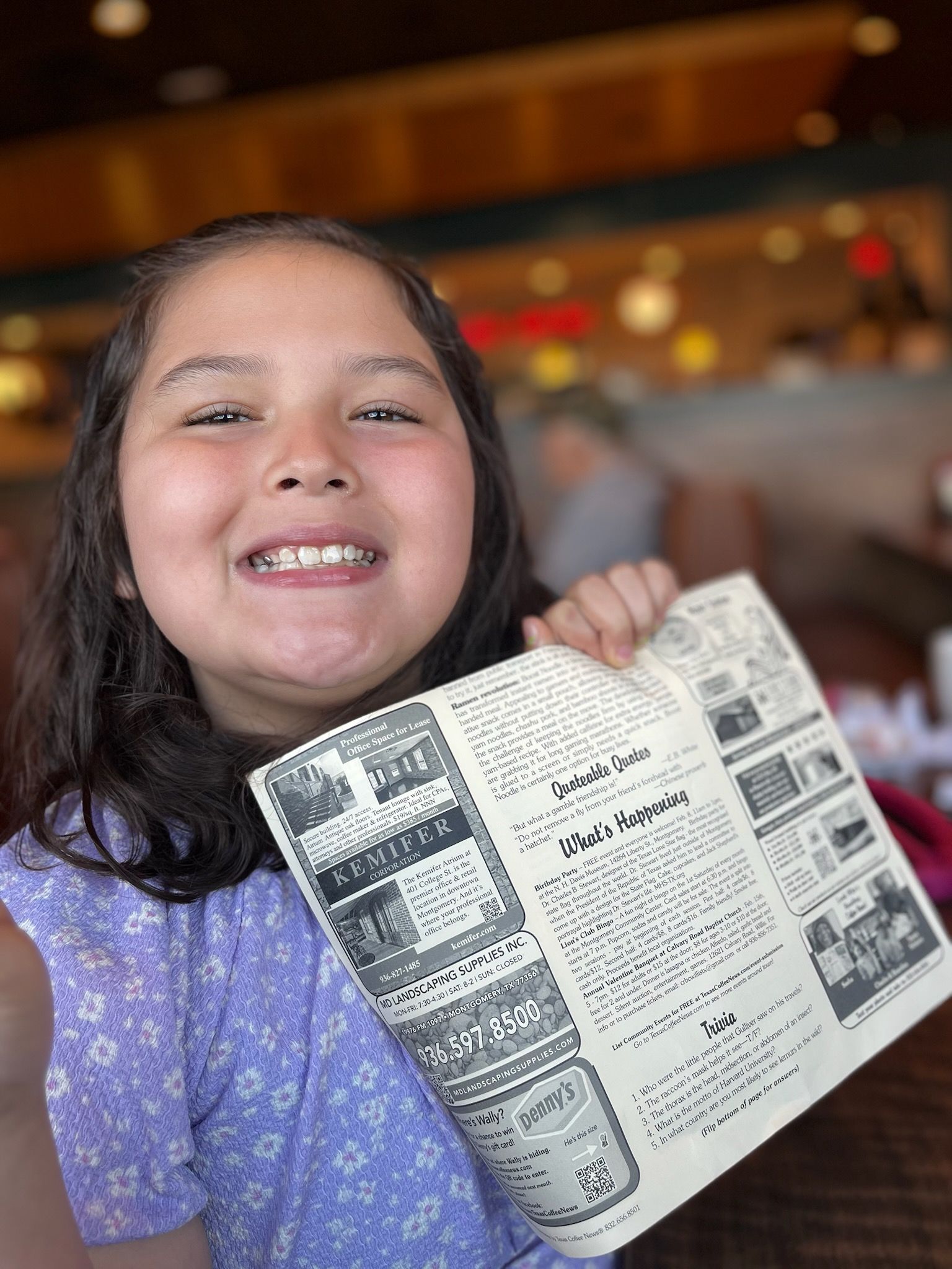 Girl with braces smiles, holding a restaurant menu. Wearing a purple floral dress, sitting in a restaurant.