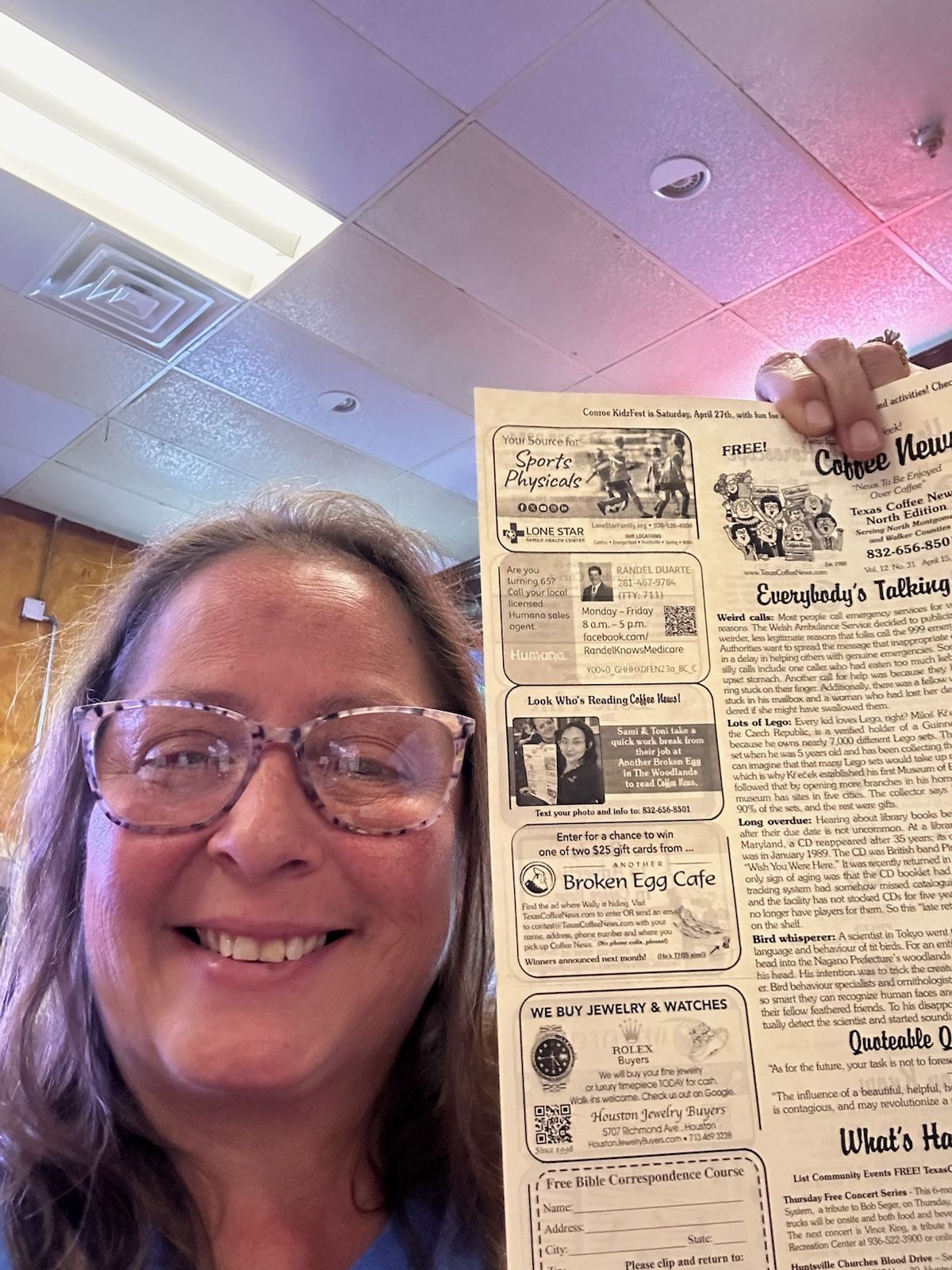 Woman holding a printed newsletter, smiling, indoor setting, overhead lighting.