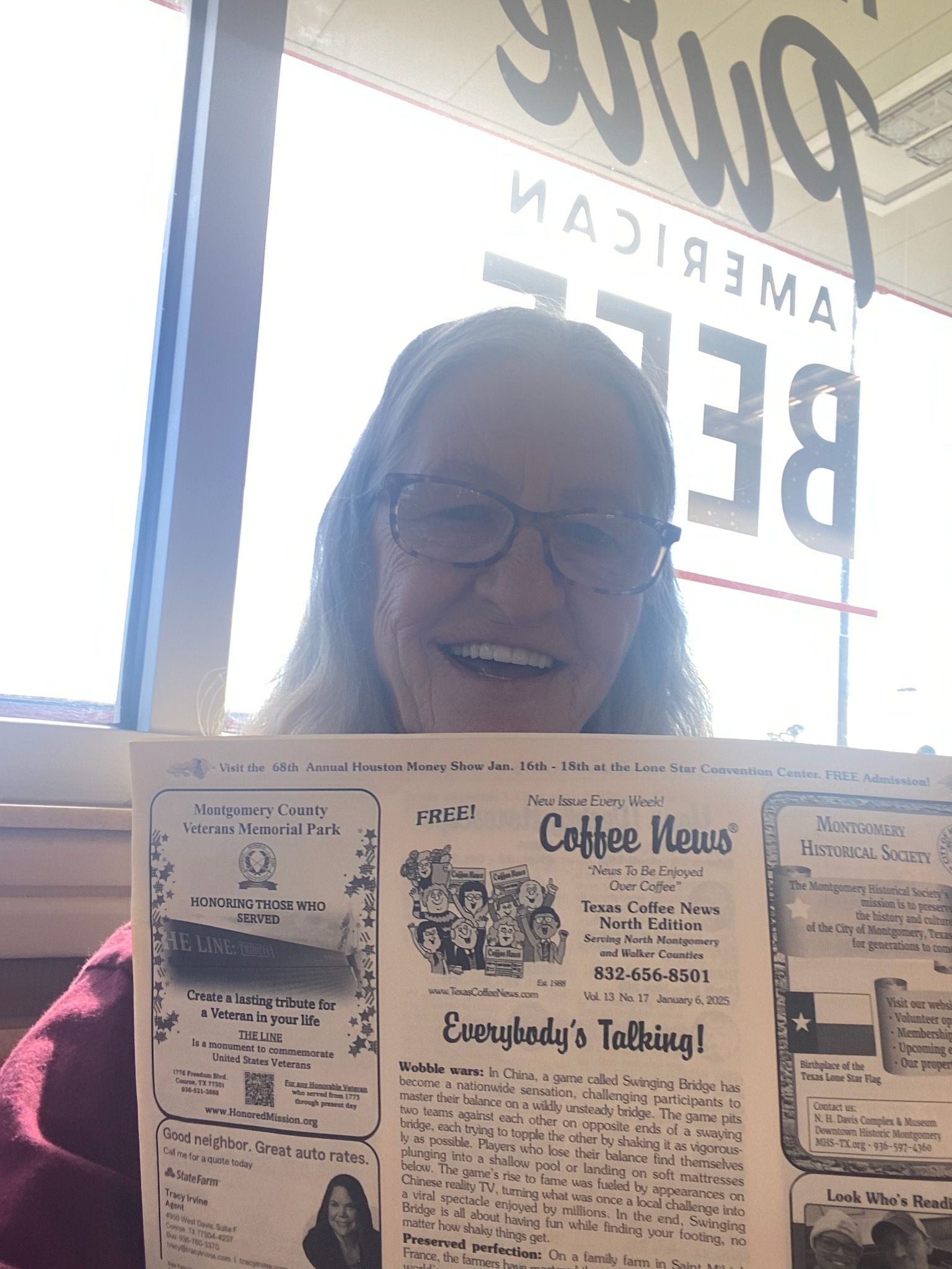 Woman holding newspaper, smiling, near window. Text in the window reads 