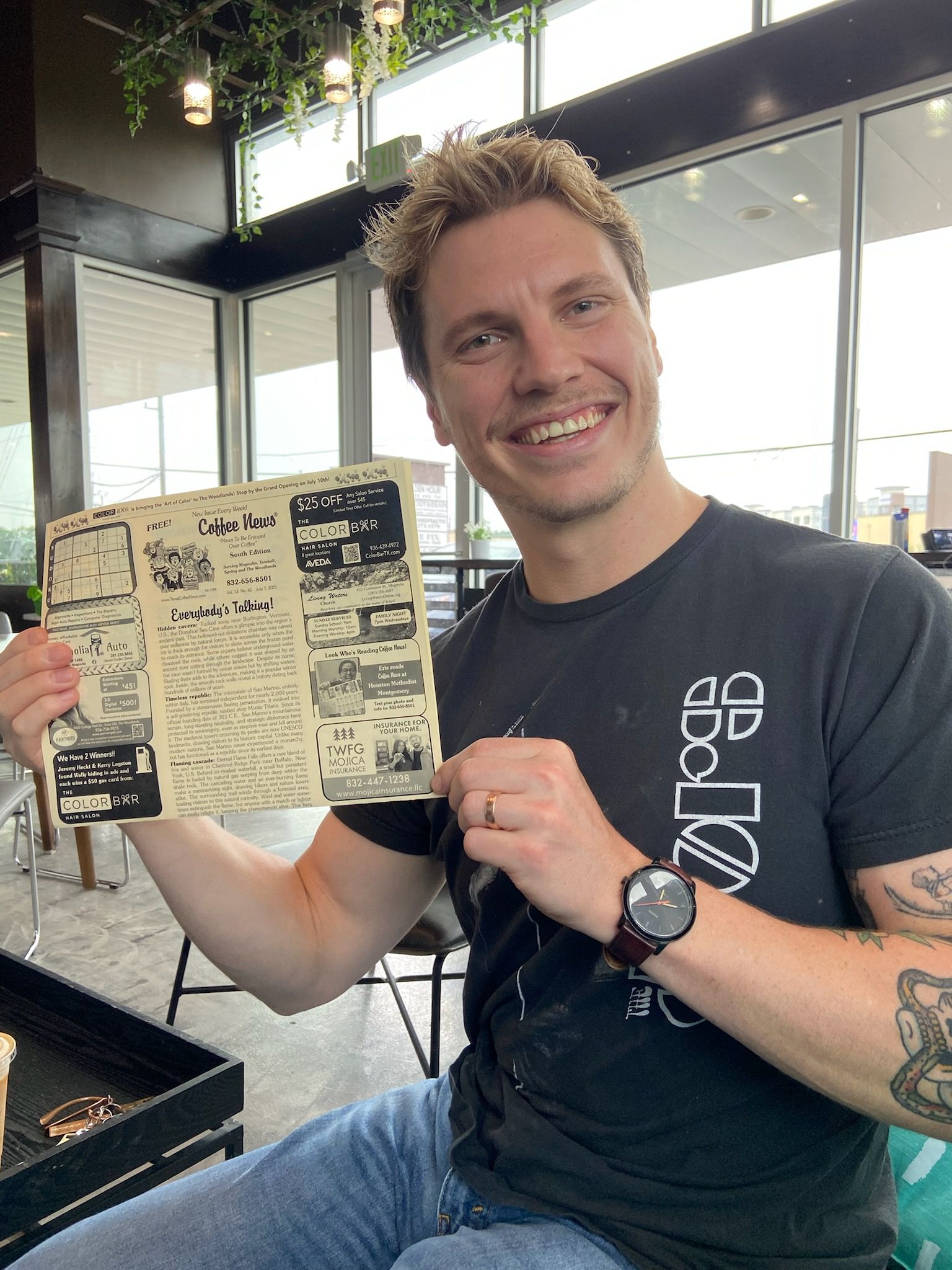 Man smiles, holding a black and white document in a bright indoor setting. He wears a black shirt, watch, and jeans.