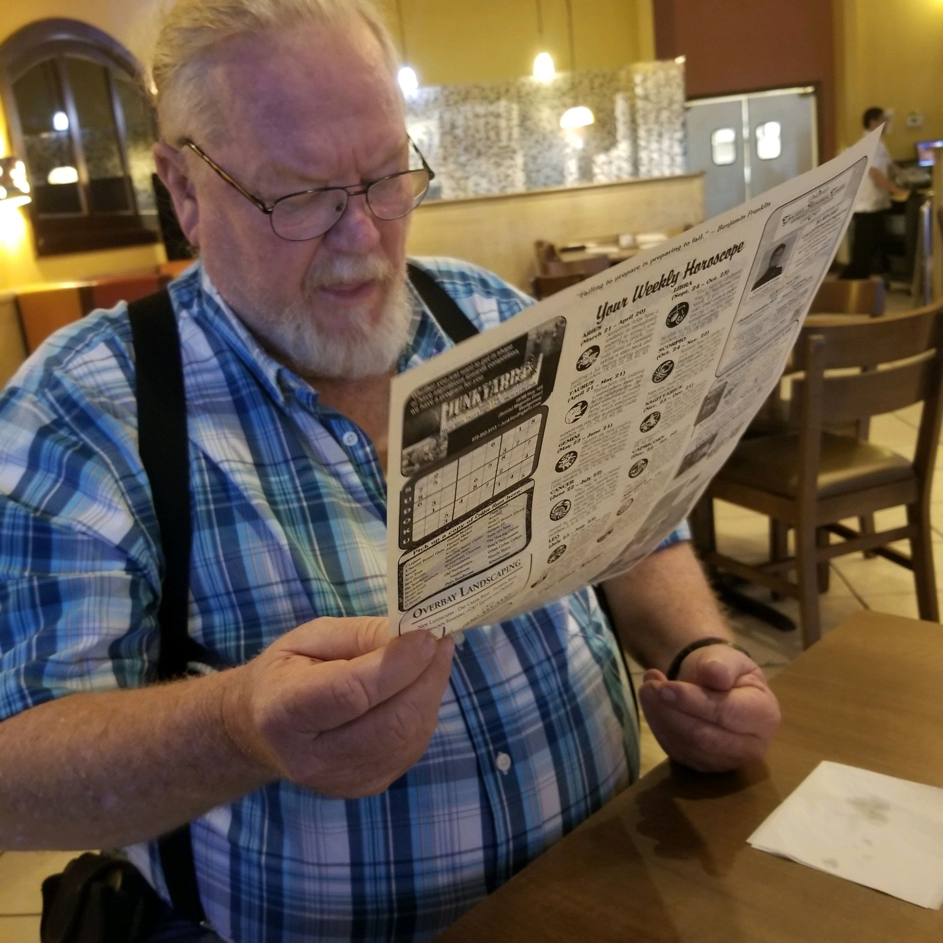 Man with glasses reading a newspaper in a restaurant, wearing a blue plaid shirt and suspenders.
