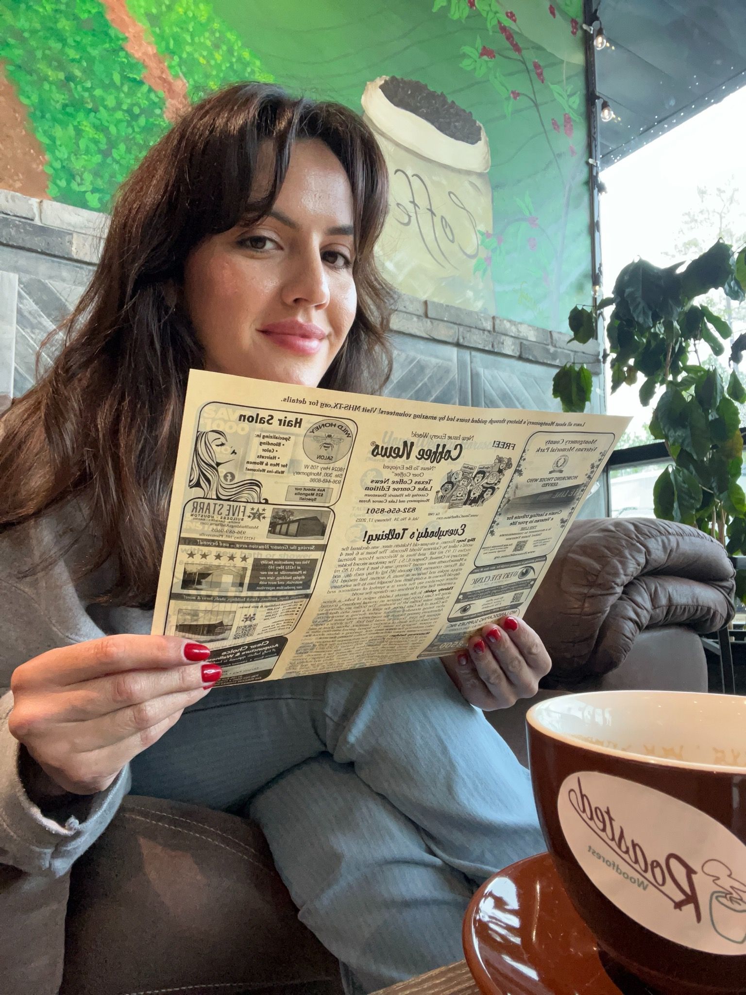 Woman in a coffee shop holding a menu, smiling. Brown mug on table, mural background.