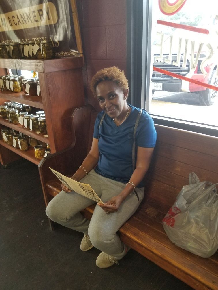 Woman sitting on a wooden bench, reading a menu, looking towards the camera. She's in a shop with jars on shelves.
