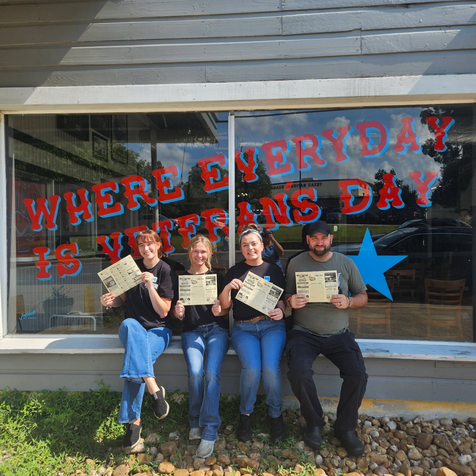 Four people holding newspapers sit in front of a window with Veterans Day messaging.