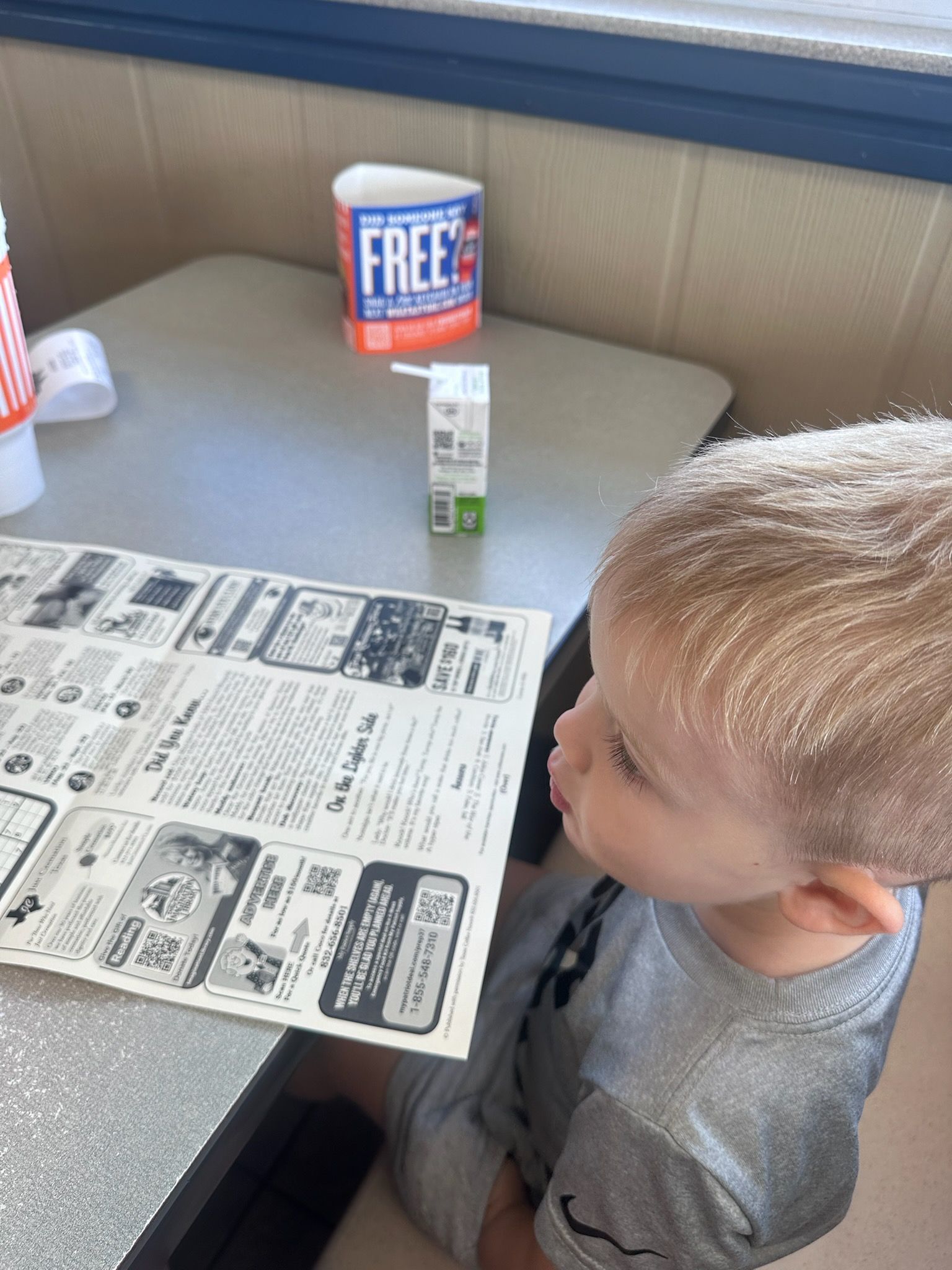 A young child looks at a newspaper on a table. A juice box and a condiment container are present.