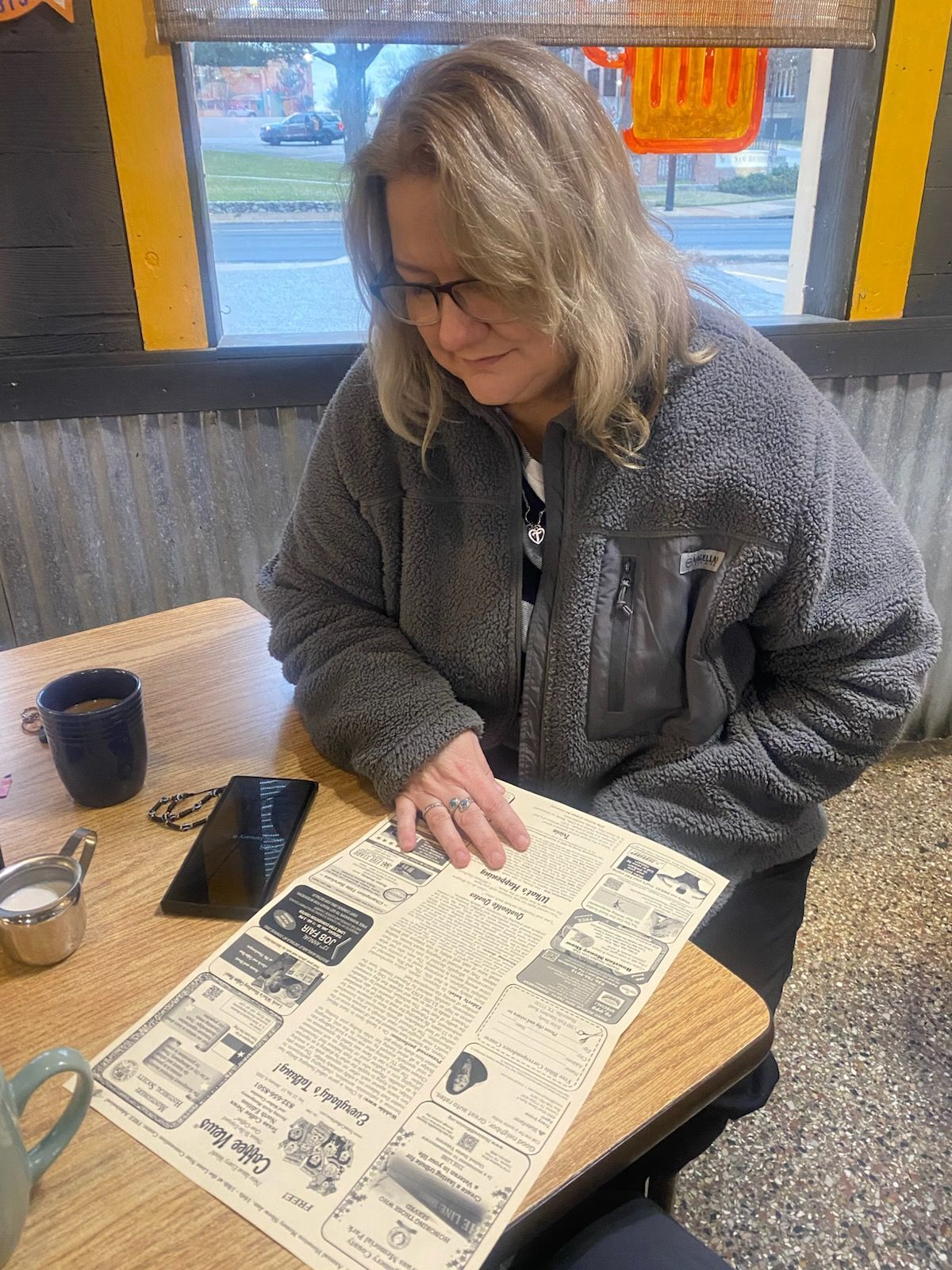 Woman in gray fleece jacket reading a menu at a table in a restaurant.