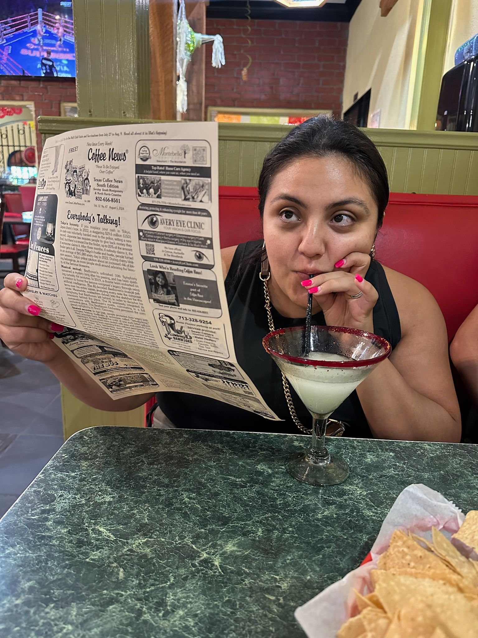 Woman in restaurant drinking from a margarita glass while holding a menu, looking up.