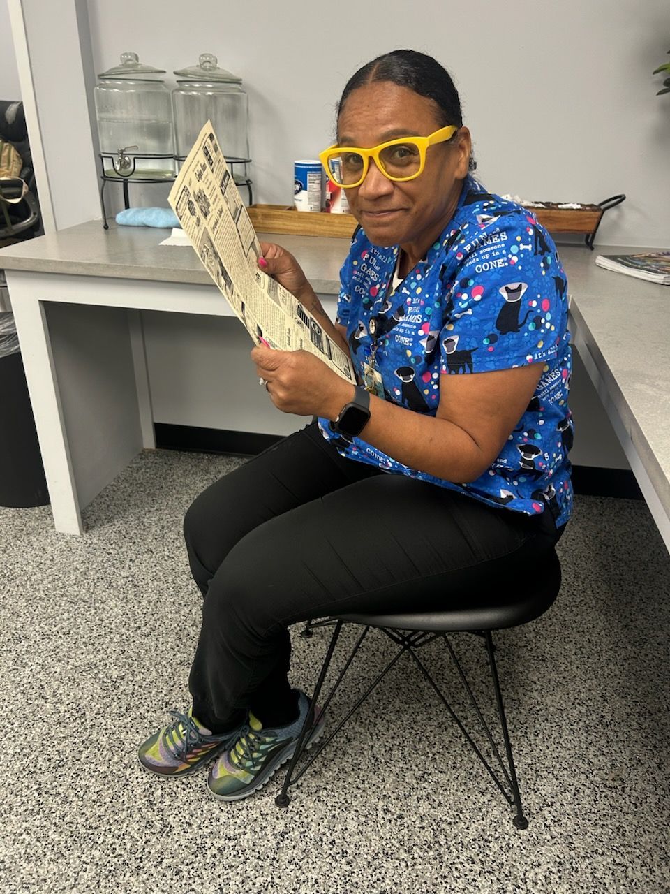 Woman in yellow glasses reads a document while seated. She wears a blue patterned shirt, black pants, and sneakers.