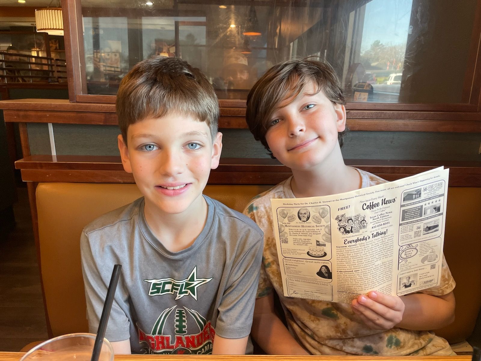 Two boys at a restaurant, one holding a menu, both smiling. Tan booth, neutral background.