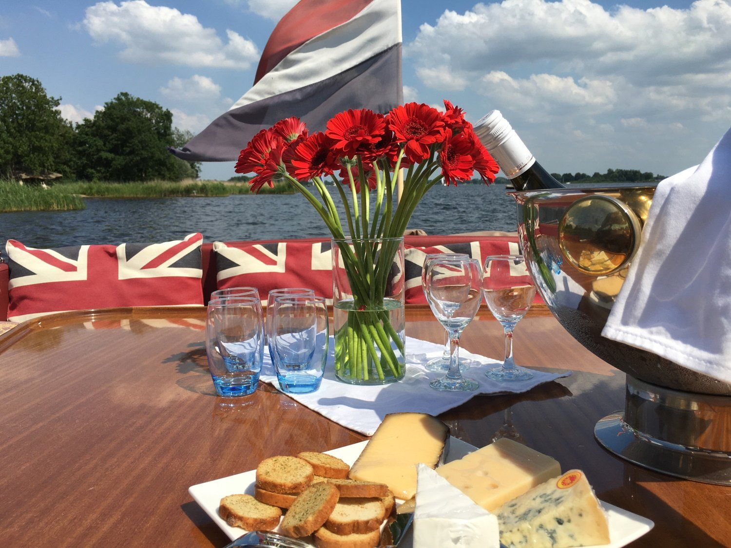 Een bootdek met rode bloemen in een vaas, een kaasplankje, crackers en een wijnkoeler tegen een schilderachtige waterachtergrond.