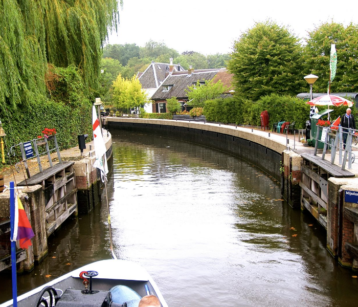 Een boot vaart richting een kleine sluis in een weelderig groen dorp met traditionele gebouwen.