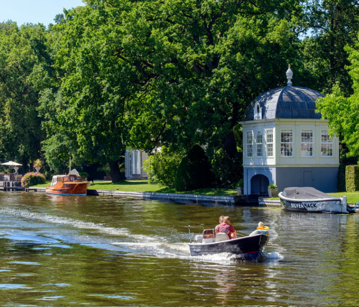 Een klein bootje vaart over een zonovergoten rivier langs een wit prieel en weelderige bomen, met andere aangemeerde boten in de buurt.