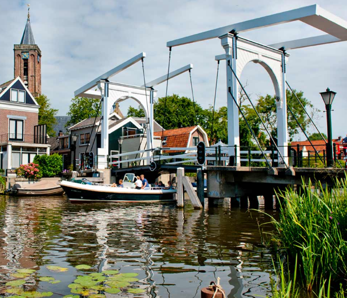 Een motorboot vaart onder een traditionele witte ophaalbrug door in een kanaal, met historische gebouwen en een kerktoren in de buurt.