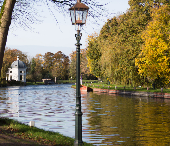 Op de voorgrond staat een straatlantaarn, met uitzicht op een rustig kanaal, en op de achtergrond een wit paviljoen en goudkleurige herfstbomen.