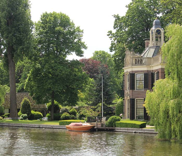Een historisch huis met een klokkentoren staat langs een kanaal, omgeven door weelderige bomen, met een klein bootje aangemeerd aan de stenen oever.