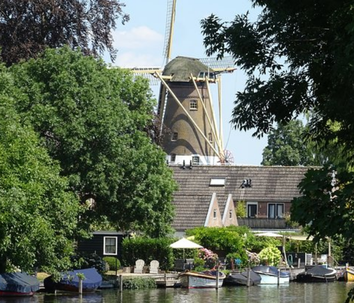 Een traditionele windmolen torent boven een tuin aan het kanaal uit, met boten aangemeerd langs de oever, omgeven door weelderige groene bomen.