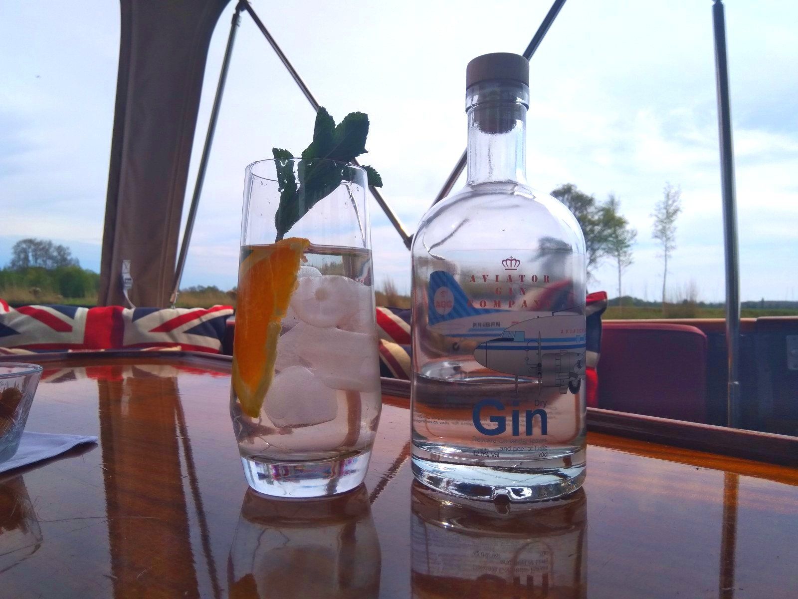 Een glas gin met een schijfje sinaasappel en munt, naast een fles gin met een luchtvaartthema, op een tafeltje buiten.