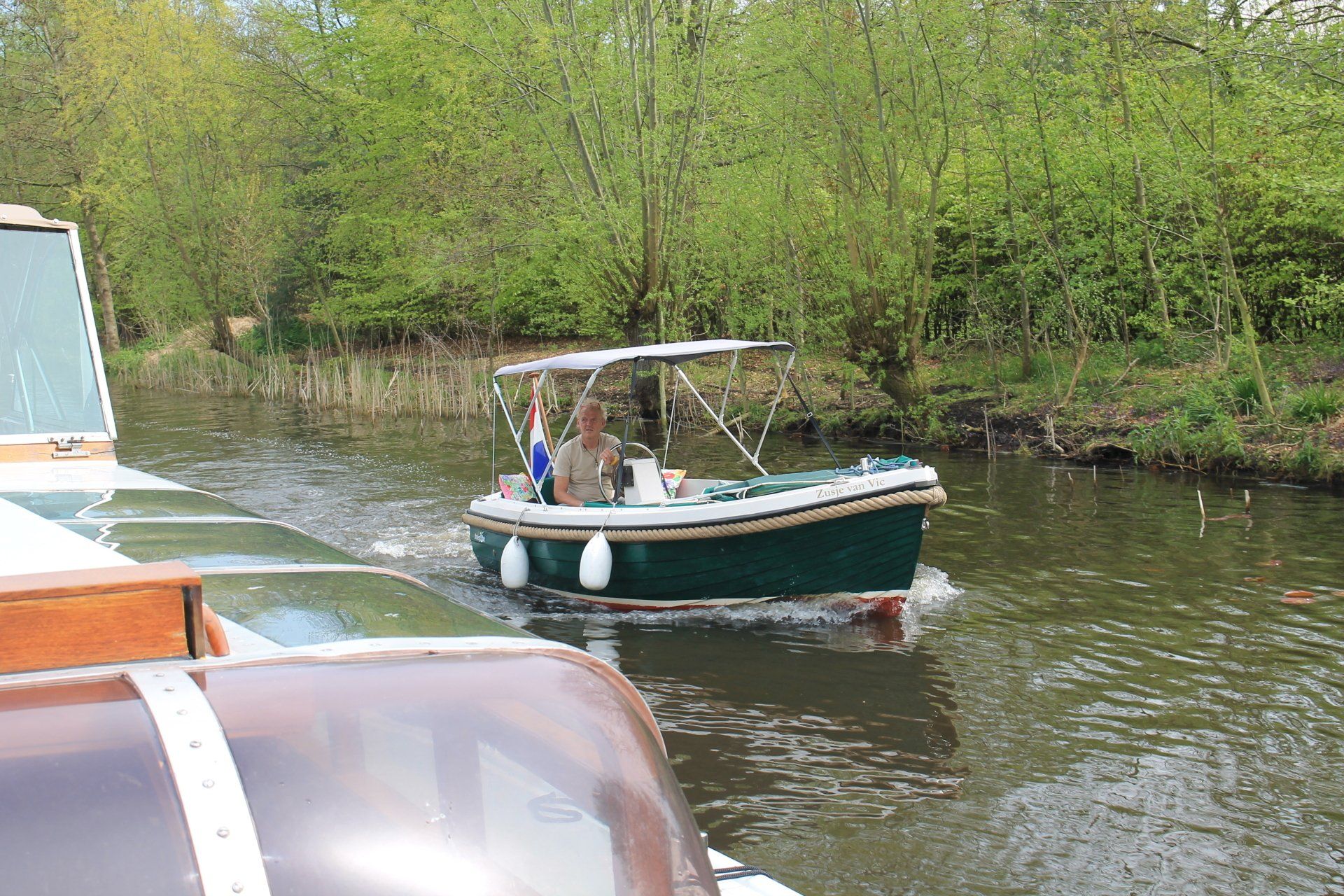 Een klein, donkergroen bootje met een overkapping vaart over een kanaal langs een beboste oever, gezien vanaf een naburige boot.