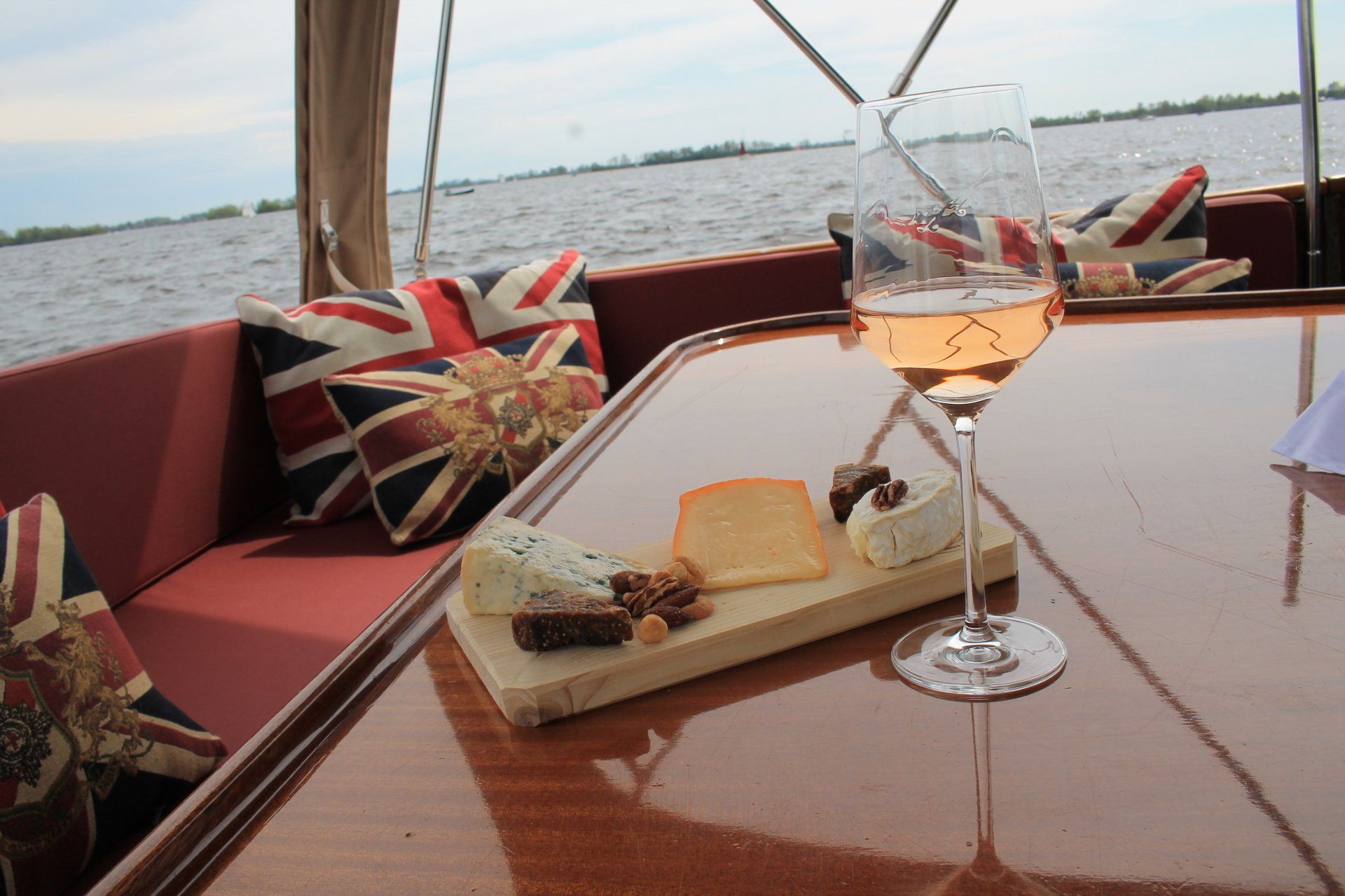 Een wijnglas en een klein kaasplankje op een houten tafel, met kussens met de Britse vlag op het dek van een boot met uitzicht op het water.