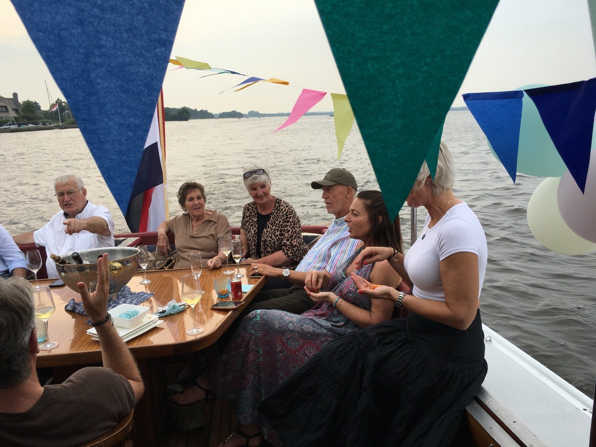 Een groep mensen zit rond een houten tafel op een boot, omringd door feestelijke driehoekige vlaggen, met uitzicht op het water op de achtergrond.