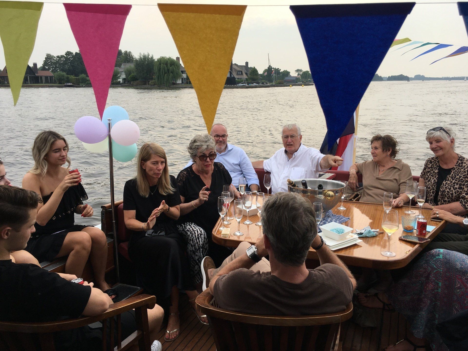 Een groep mensen zit rond een tafel op een boot die versierd is met kleurrijke vlaggetjes, en geniet overdag van een drankje.