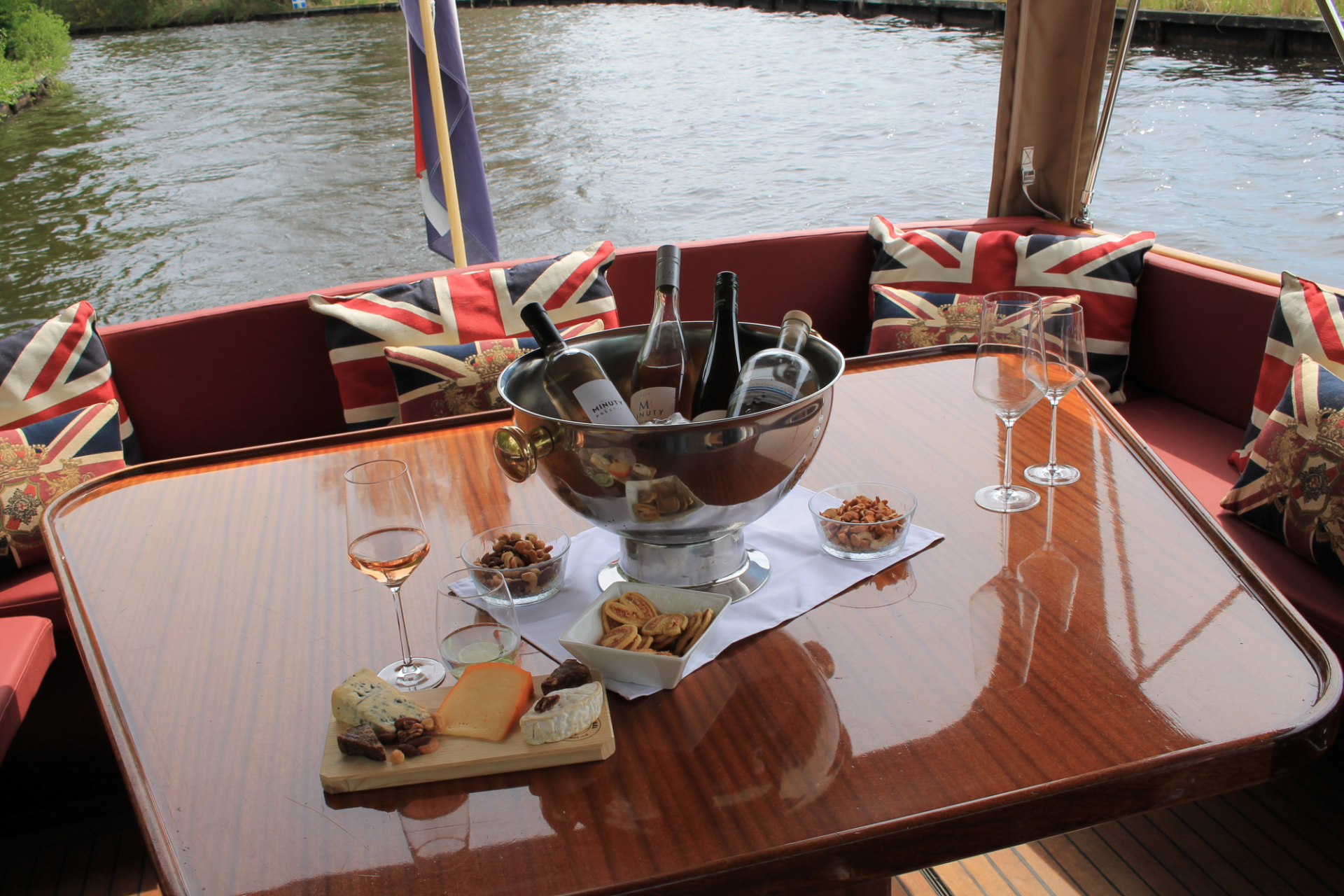 Een wijnkoeler met flessen, een kaasplank en wijnglazen op een gepolijste houten tafel op een boot met kussens met de Britse vlag.