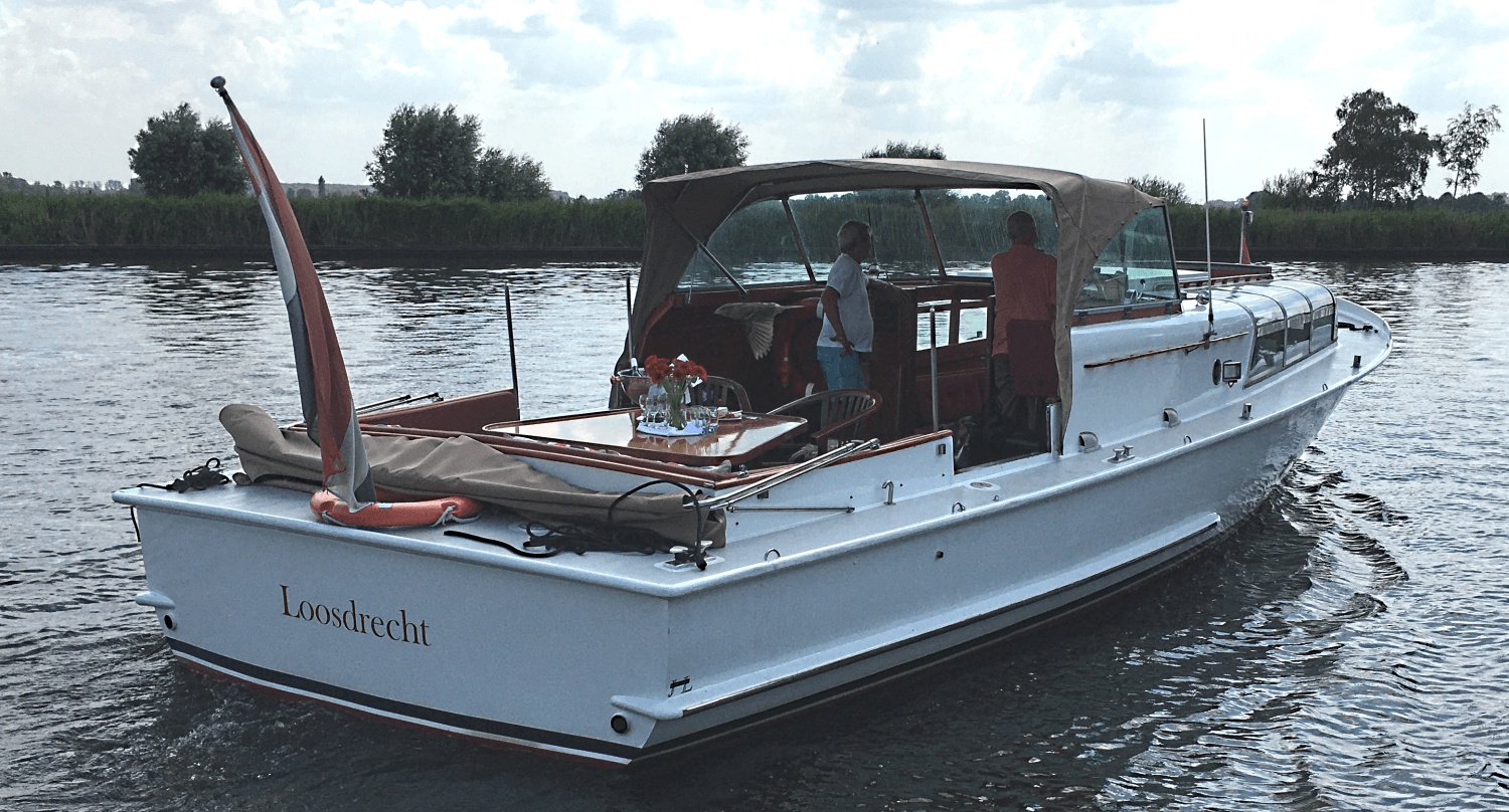 Een witte motorboot met een beige canvas overkapping vaart op een bewolkte dag over een rivier, met twee mensen aan dek.