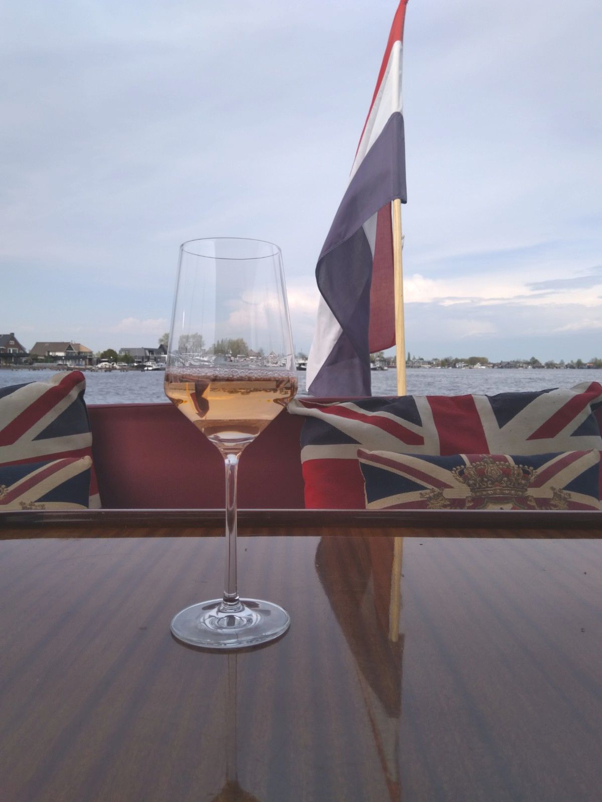 Een wijnglas gevuld met rosé staat op een houten tafel op een boot, met kussens waarop de Britse vlag en een Nederlandse vlag zijn afgebeeld.