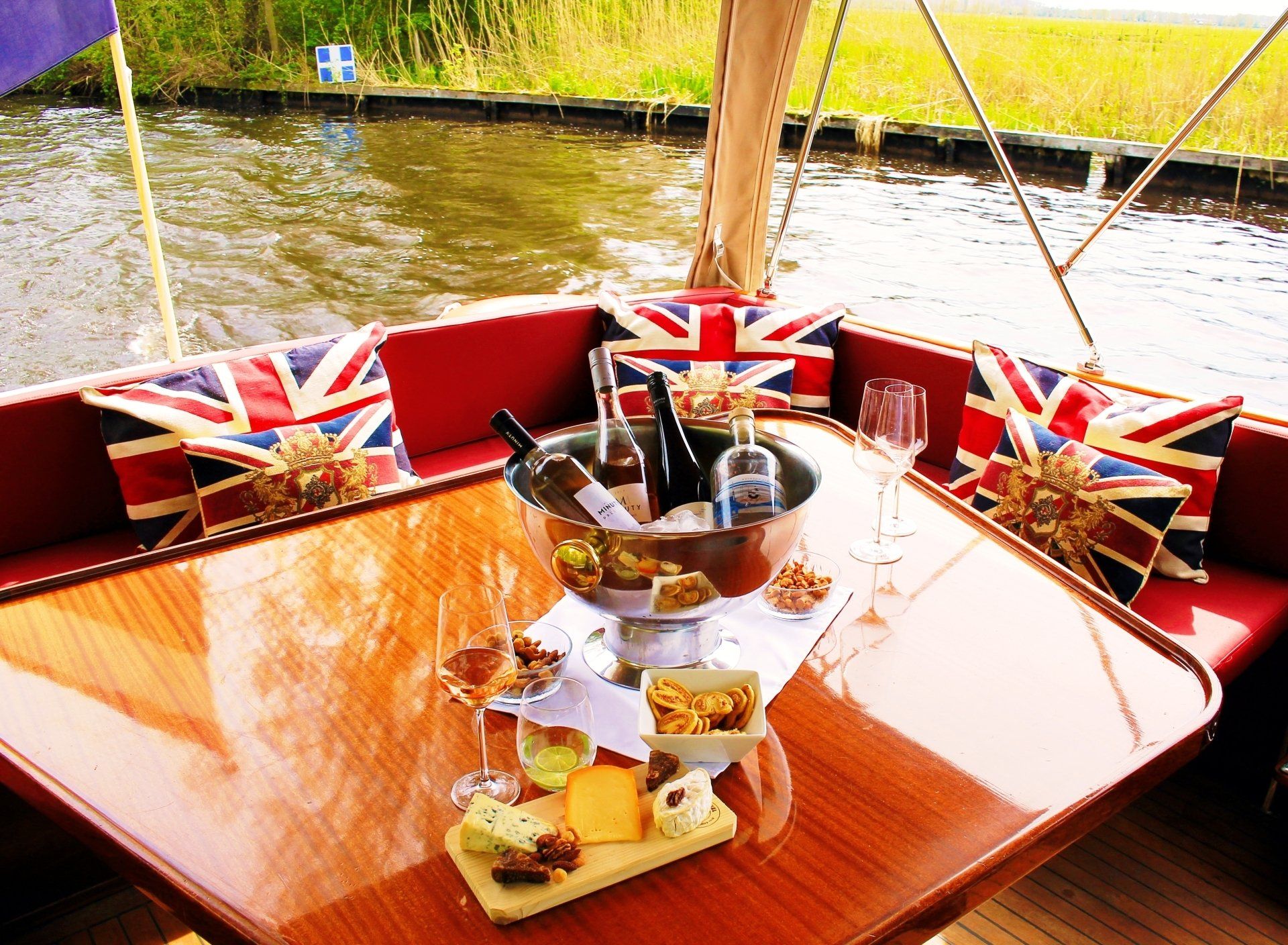 Een tafel met wijn, kaas en hapjes, geplaatst op een boot met kussens in de vorm van de Britse vlag, met uitzicht op een rivier.