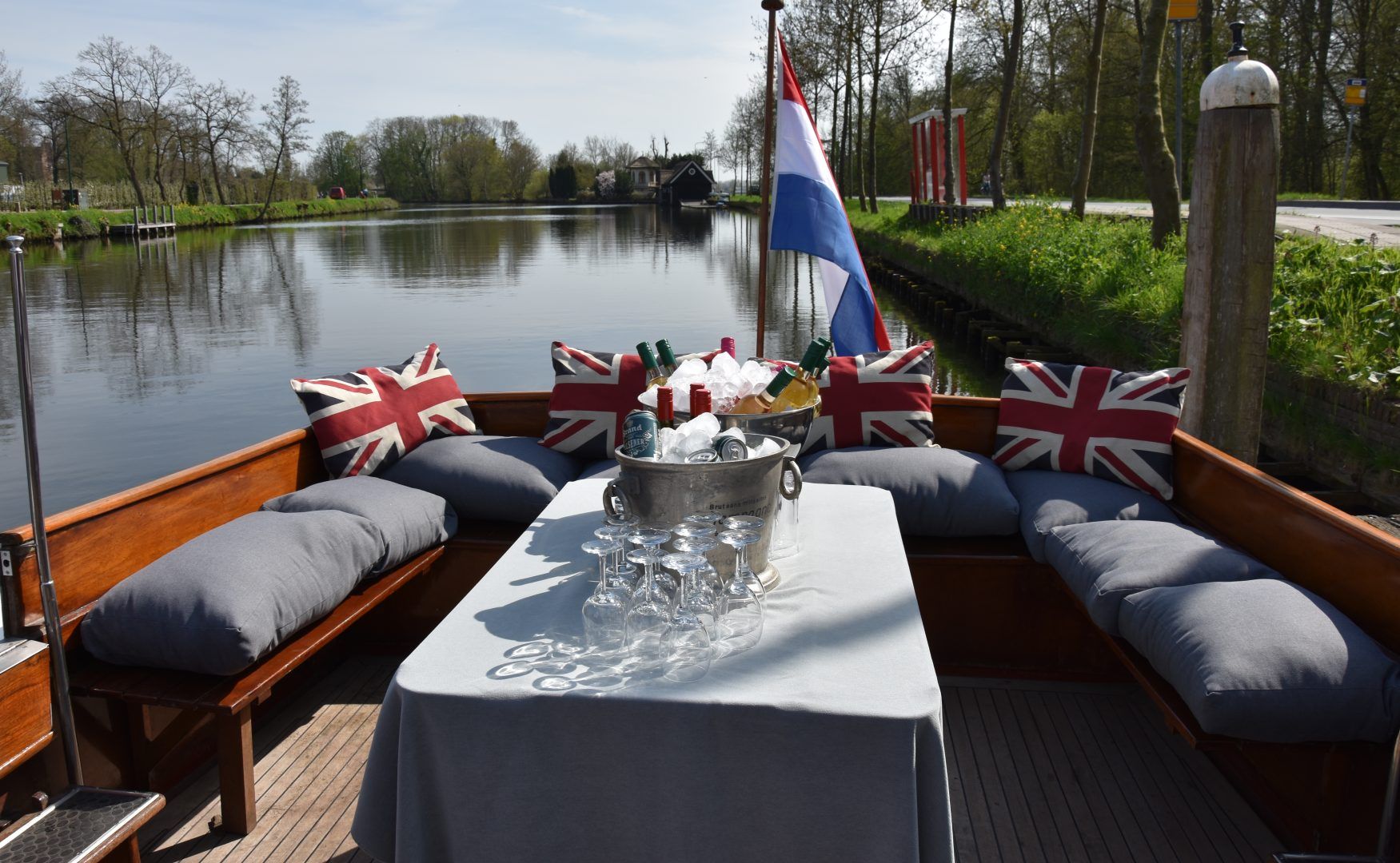 Een bootdek op een kanaal met een tafel, een champagnekoeler en een U-vormige zithoek met kussens in de kleuren van de Britse vlag en een Nederlandse vlag.