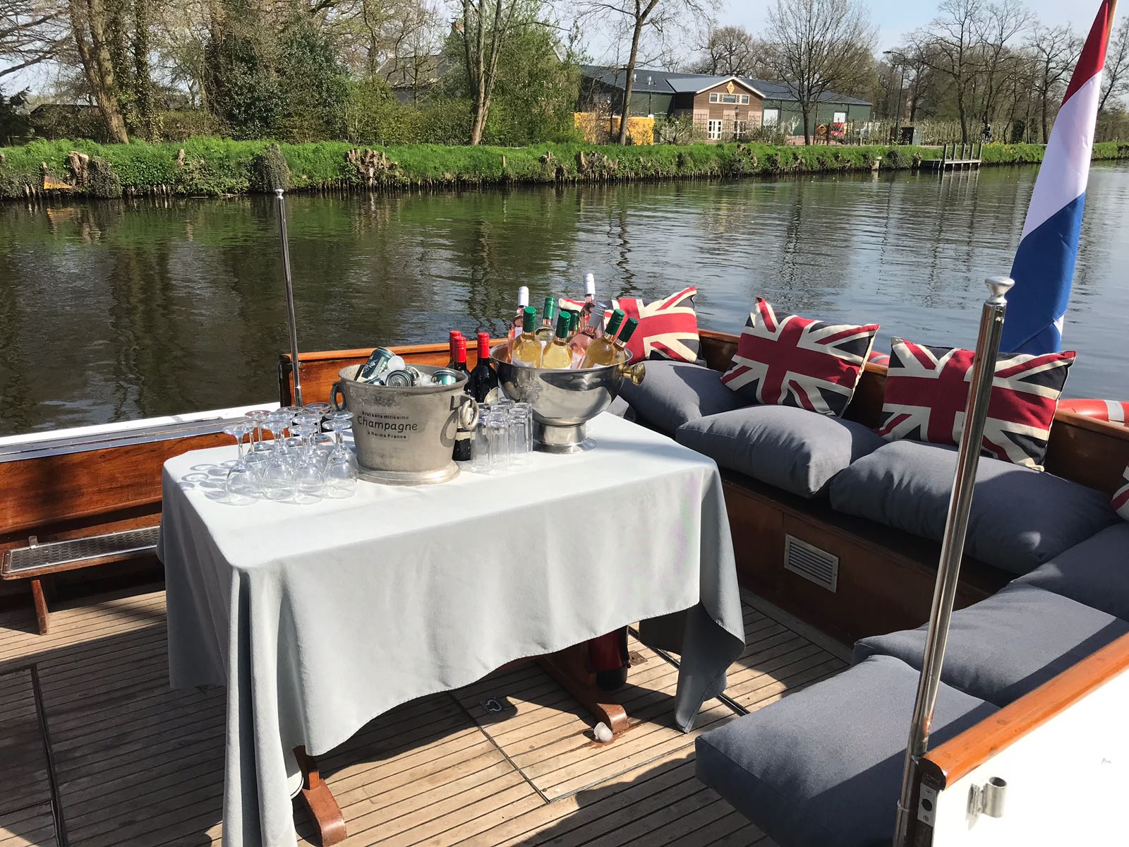 Een tafel met wijnkoelers en glazen op het dek van een boot, met comfortabele zitplaatsen voorzien van kussens met de Britse vlag.