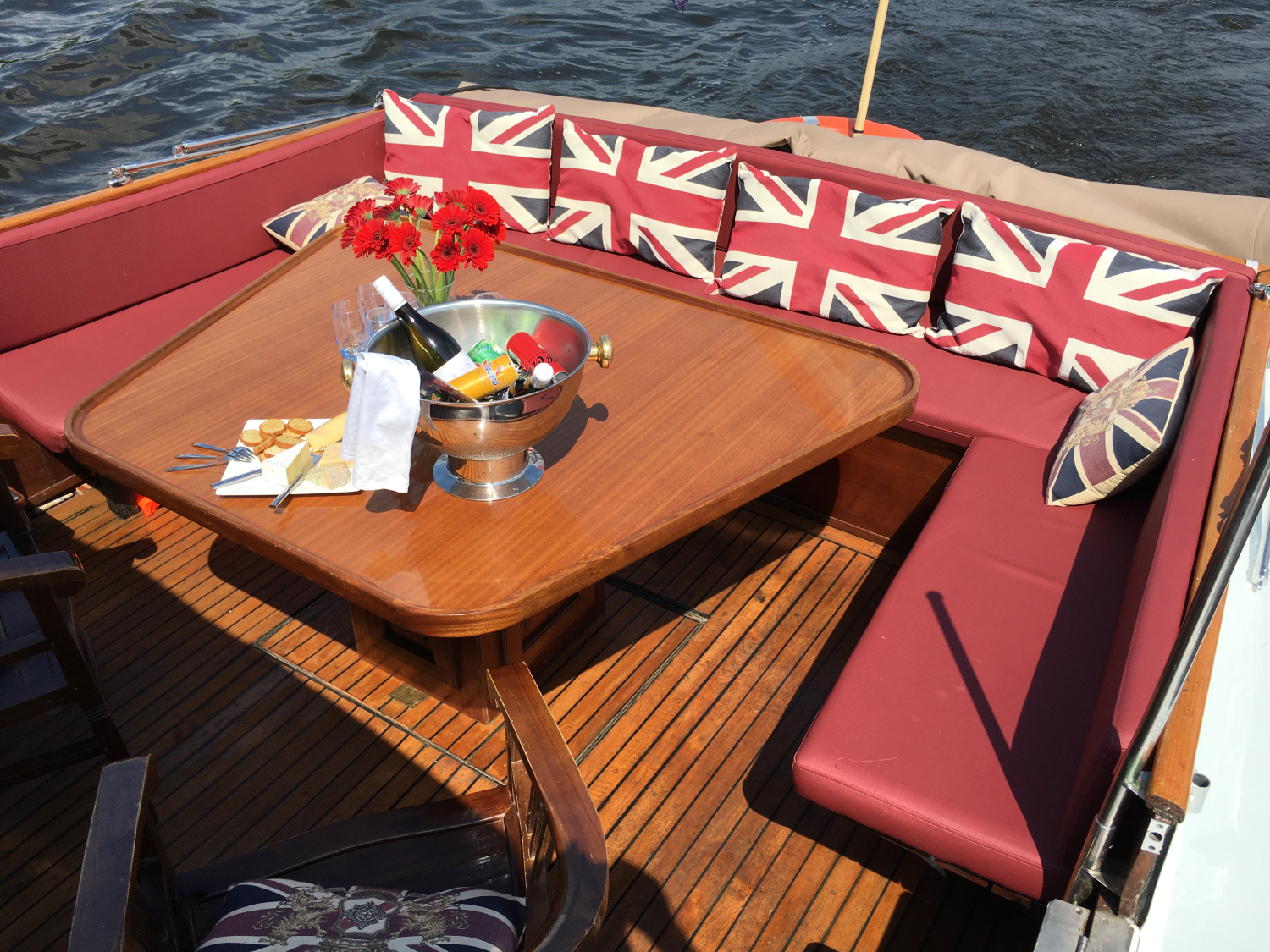 Een houten bootdek met rode zitbanken, kussens met de Britse vlag, een eettafel, bloemen en een ijsemmer met drankjes.