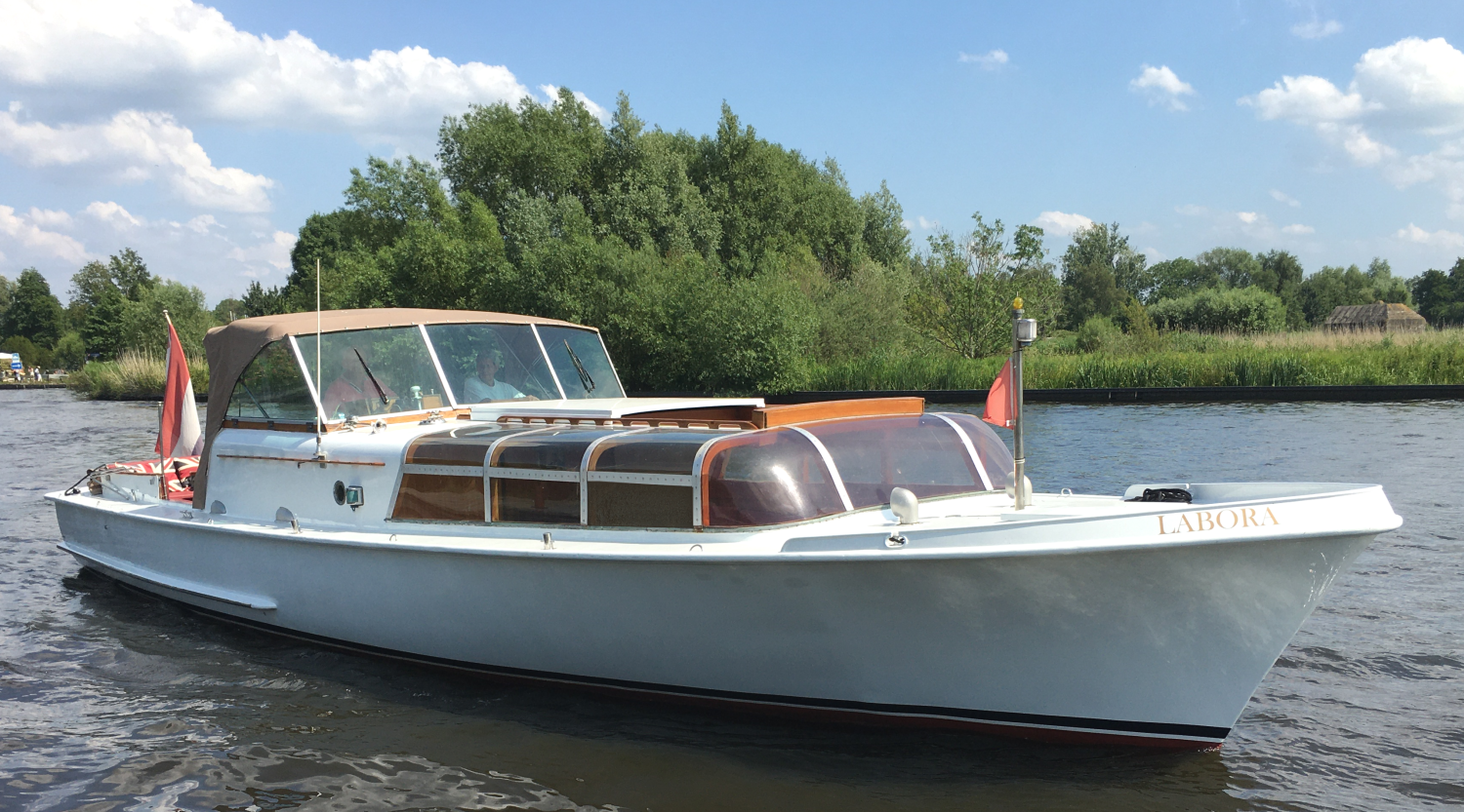 Een witte, klassieke motorboot vaart rustig over een rivier, met op de achtergrond groene bomen onder een blauwe hemel met wolken.