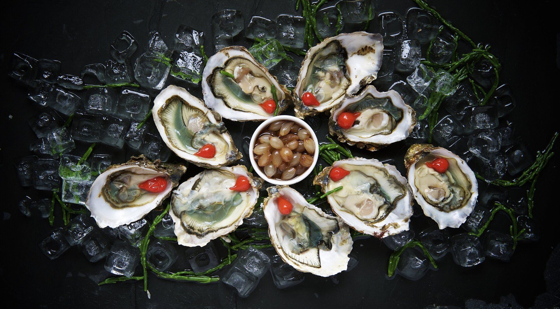 Acht oesters, gerangschikt op een bedje van ijs rond een centrale schaal met kleine bruine bessen, elk bedekt met een rode saus.