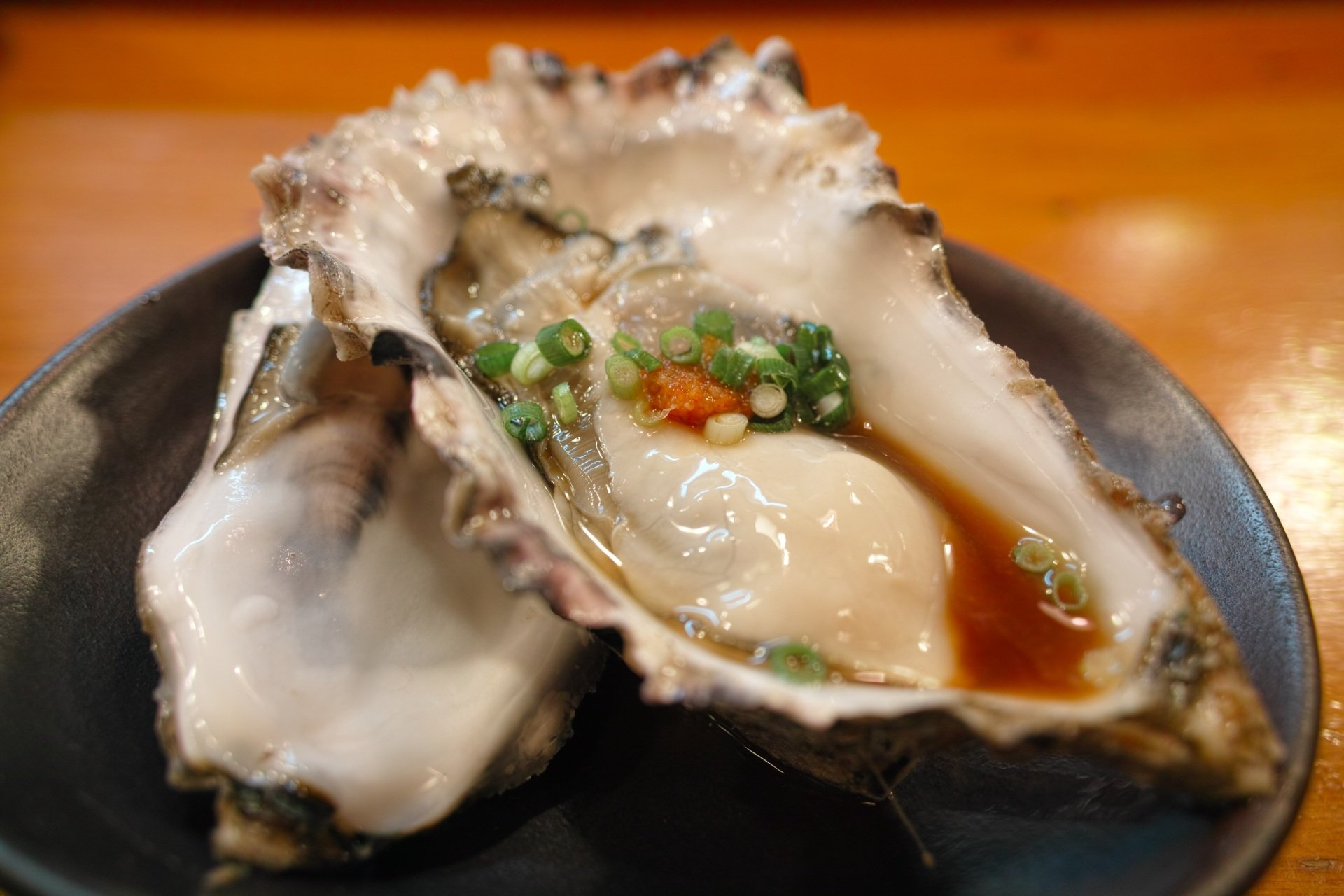 Twee rauwe oesters op een donker bord, gegarneerd met lente-uitjes en een klodder chilisaus in een ondiepe, hartige vloeistof.