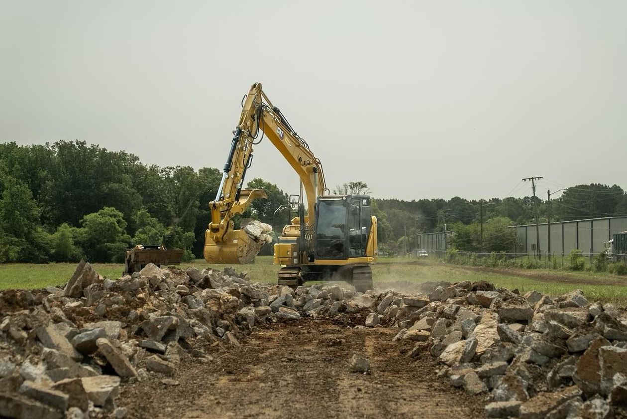 Yellow excavator demolishing concrete debris in a field, trees in the background.