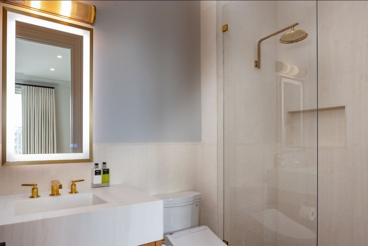 A modern bathroom with a gold-trimmed mirror, white vanity, gold faucet, and a glass-enclosed shower with gold fixtures.