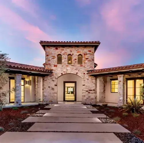 A stone-faced Mediterranean home with a tile roof, centered front door, and walkway set against a vibrant sunset sky.
