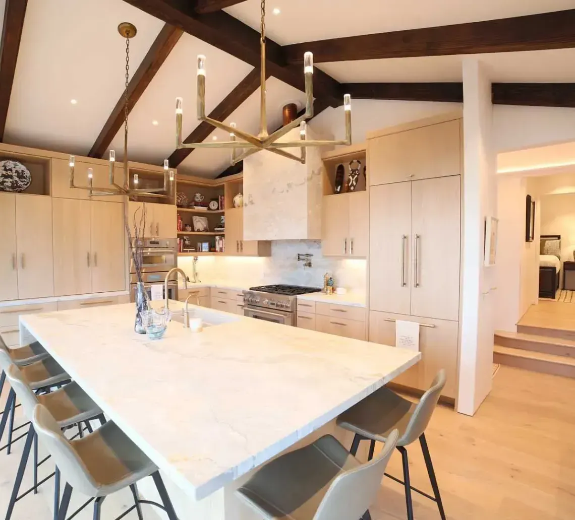 A modern kitchen with light wood cabinets, a large stone island, bar seating, and exposed dark ceiling beams.