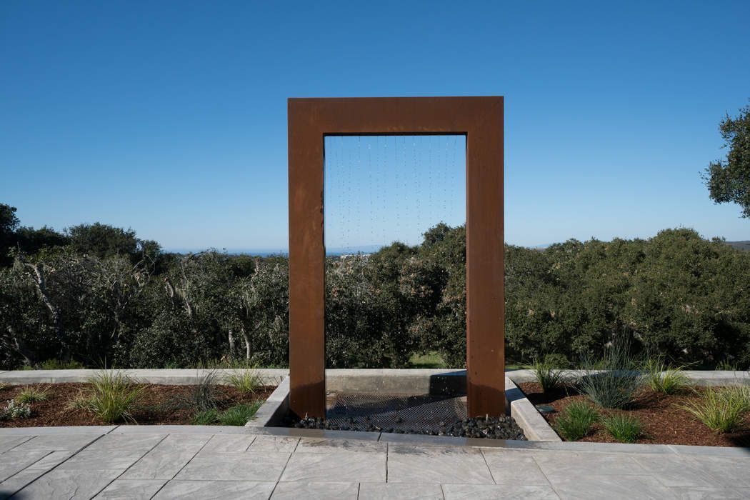 A rust-colored rectangular metal frame stands on a stone patio overlooking a tree-covered landscape under a clear sky.