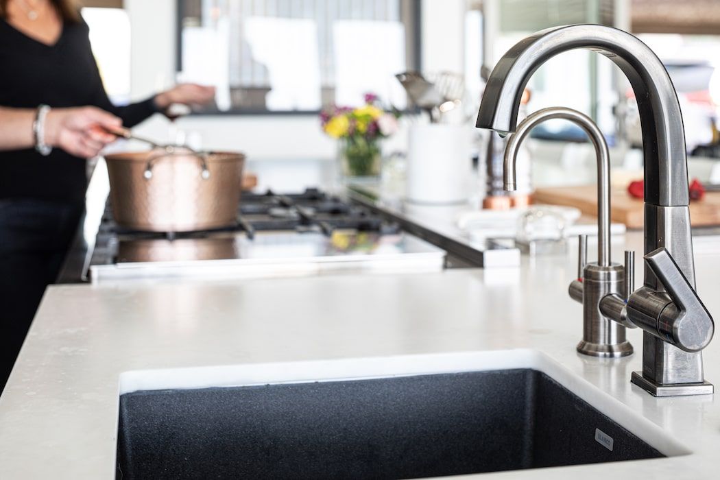A modern kitchen island with a dark sink and metallic faucet in the foreground as someone cooks on a stove behind it.