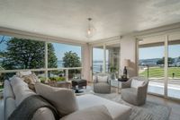 A sunlit living room with a cream sofa, two armchairs, and floor-to-ceiling windows overlooking a coastal view and lawn.