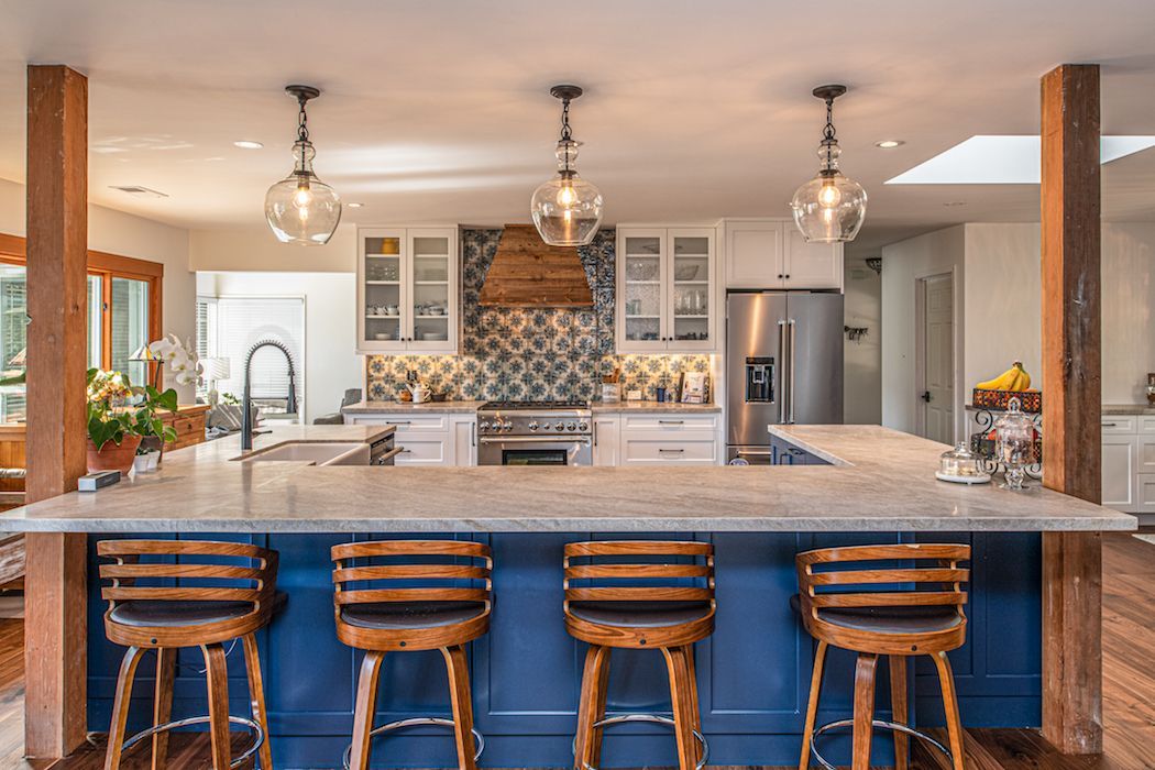 A kitchen featuring a blue island with four wooden stools, white cabinets, a patterned backsplash, and three pendants.